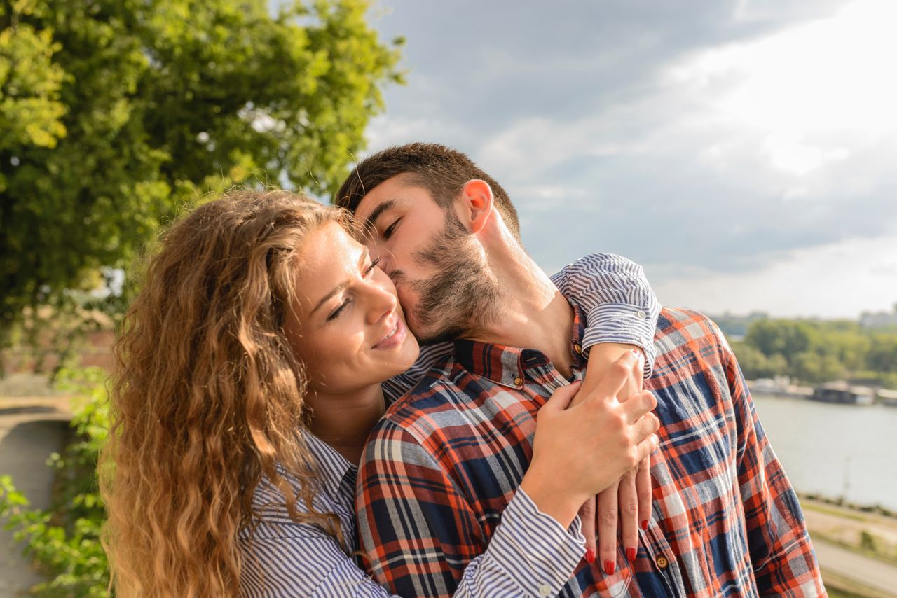 A woman hugs her partner from behind, while he leans back and kisses her on the cheek.